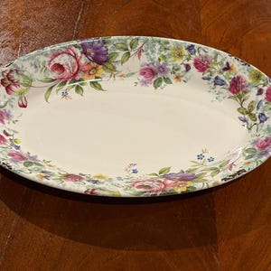 May include: Oval serving platter with a floral design. The platter features a cream-colored center and a border adorned with pink, purple, and yellow flowers, along with green foliage. The rim has a scalloped edge.