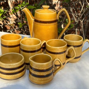 Puede incluir: Juego de té de cerámica vintage en tonos amarillos y marrones. El juego incluye una tetera, seis tazas, una jarra de crema y un azucarero. Cada pieza presenta una banda marrón con un diseño decorativo.