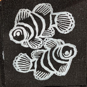 May include: A square black fabric patch featuring an embroidered design of two clownfish in light grey thread. The fish are positioned one above the other, showcasing detailed fin and stripe patterns stitched onto the dark textile surface.