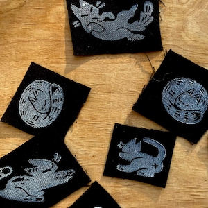 Könnte beinhalten: Eine Sammlung schwarzer Stoffflicken mit silbernen Katzenmotiven. Die Katzenillustrationen zeigen verschiedene Posen, wie eingerollte und verspielte Positionen. Die quadratischen Flicken sind auf einer Holzoberfläche angeordnet.