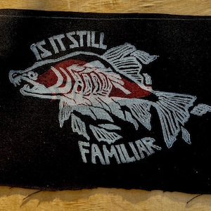 以下が含まれることがあります： 赤と白のアクセントが施された、黒い長方形のパッチ。魚のイラストの上と下に「IS IT STILL FAMILIAR」の文字がプリントされています。衣類やアクセサリー用のパッチです。