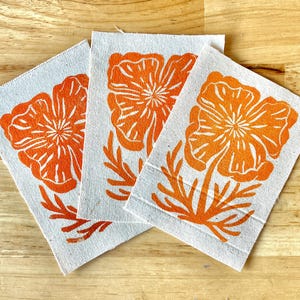 May include: Three square fabric patches with orange poppy flower designs. The flowers are centered on the patches, with detailed petals and stems. The patches are on a light-colored wooden surface.