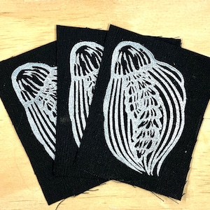 May include: Three rectangular black fabric patches, each featuring a white screen-printed graphic of a stylized seashell. The patches are arranged in a fan-like overlapping pattern, displaying the same detailed shell illustration on each piece.