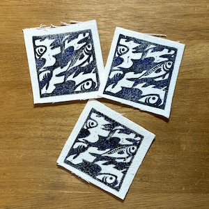May include: Three square fabric patches with a blue and white fish design. The design features stylized fish and eye shapes. The patches are arranged on a wooden surface.
