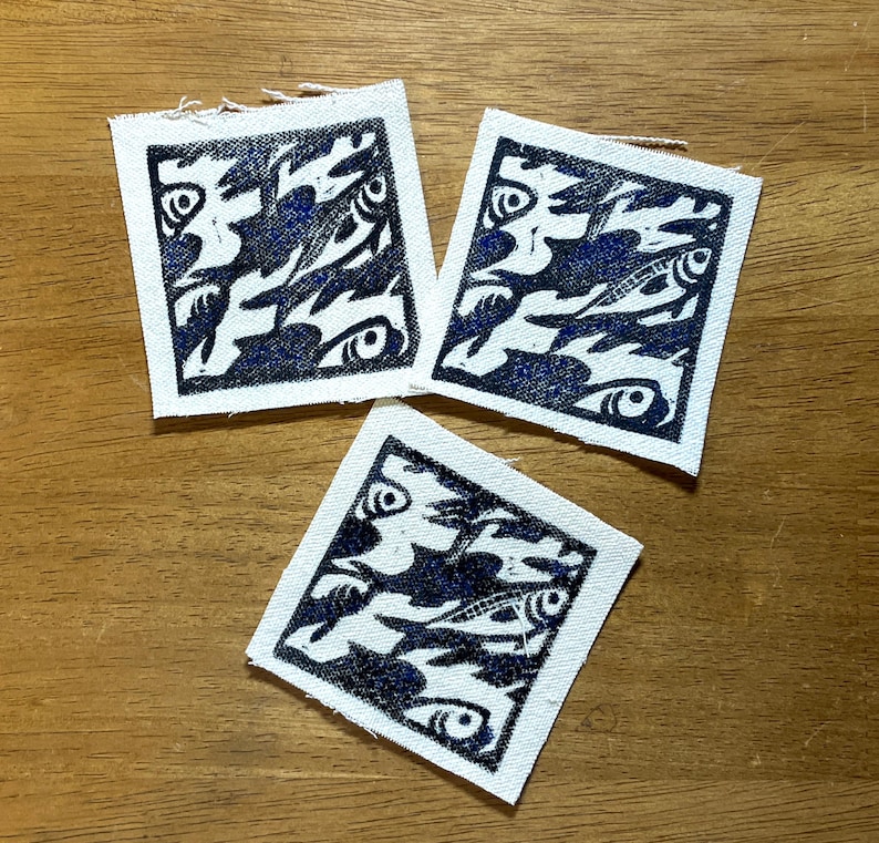 May include: Three square fabric patches with a blue and white fish design. The design features stylized fish and eye shapes. The patches are arranged on a wooden surface.