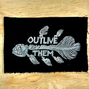 Puede incluir: Parche de tela negra con un diseño plateado de esqueleto de tortuga y las palabras "OUTLIVE THEM" en letras plateadas. El parche tiene un borde sin rematar.