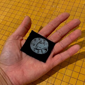 May include: A small, square black patch with a light blue embroidered design of a sleeping cat inside a circle. The patch is held in a hand against a yellow grid background.