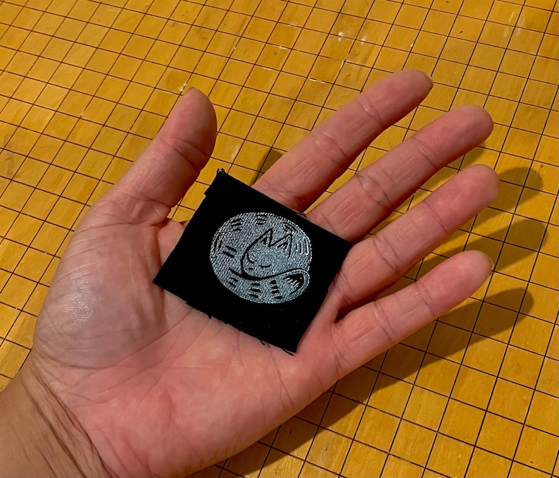 May include: A small, square black patch with a light blue embroidered design of a sleeping cat inside a circle. The patch is held in a hand against a yellow grid background.