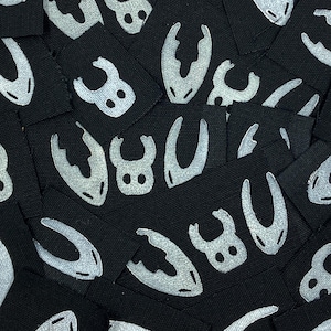 Puede incluir: Una colección de etiquetas de tela negra con un diseño blanco estilizado de garra y ojo. Las etiquetas son cuadradas y están dispersas, creando un patrón texturizado. El diseño se repite en las etiquetas.