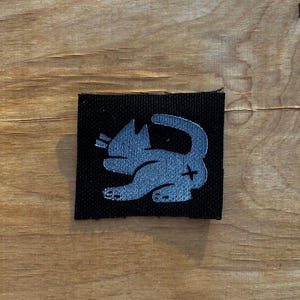 May include: A black fabric patch featuring a stylized, light blue cat design. The cat is depicted in a playful pose with its tail curled. The patch is square and appears to be made of a woven material.