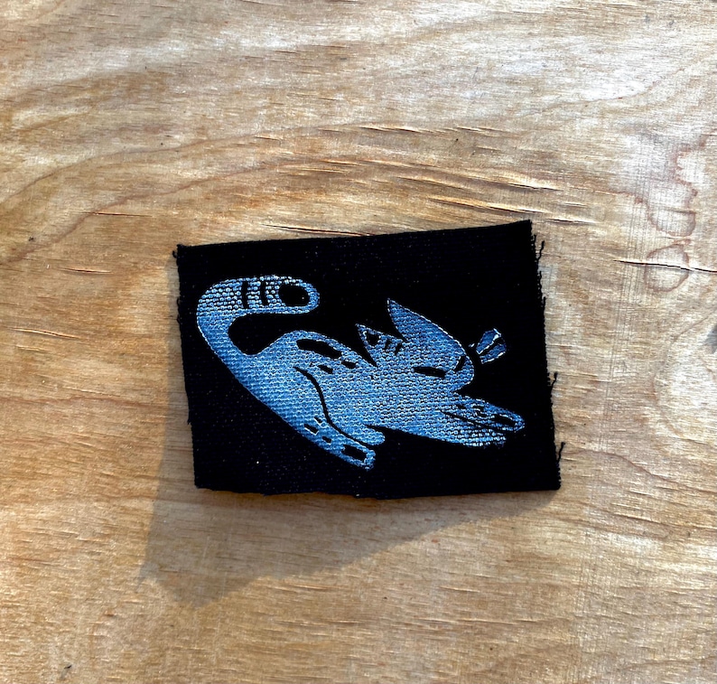 May include: A small, black fabric patch featuring a stylized, light blue illustration of a creature with a long neck and wings. The design is embroidered onto the patch, which is rectangular in shape.