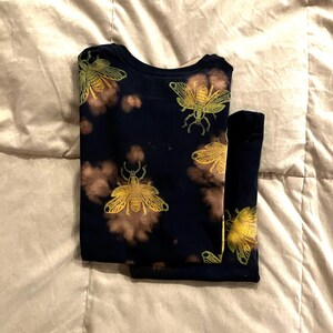 Op de afbeelding: Donker marineblauw shirt met lange mouwen en broek met een herhalend patroon van goudkleurige insectenillustraties. Het shirt en de broek hebben bruine accenten in tie-dye-stijl. De set is netjes op een licht oppervlak gevouwen.