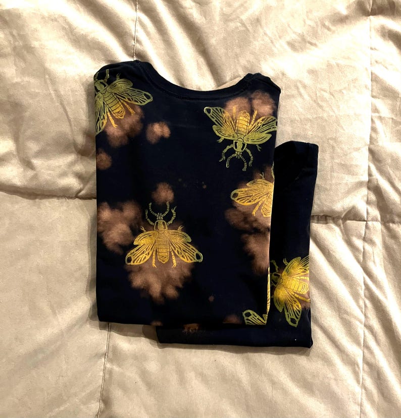 Op de afbeelding: Donker marineblauw shirt met lange mouwen en broek met een herhalend patroon van goudkleurige insectenillustraties. Het shirt en de broek hebben bruine accenten in tie-dye-stijl. De set is netjes op een licht oppervlak gevouwen.