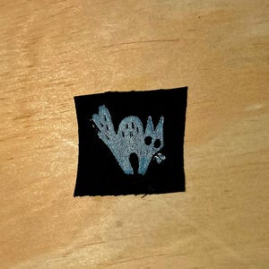 May include: A black square patch featuring a stylized, light gray cat illustration. The cat has large eyes and a curved tail, with a slightly open mouth. The patch is likely for clothing or accessories.