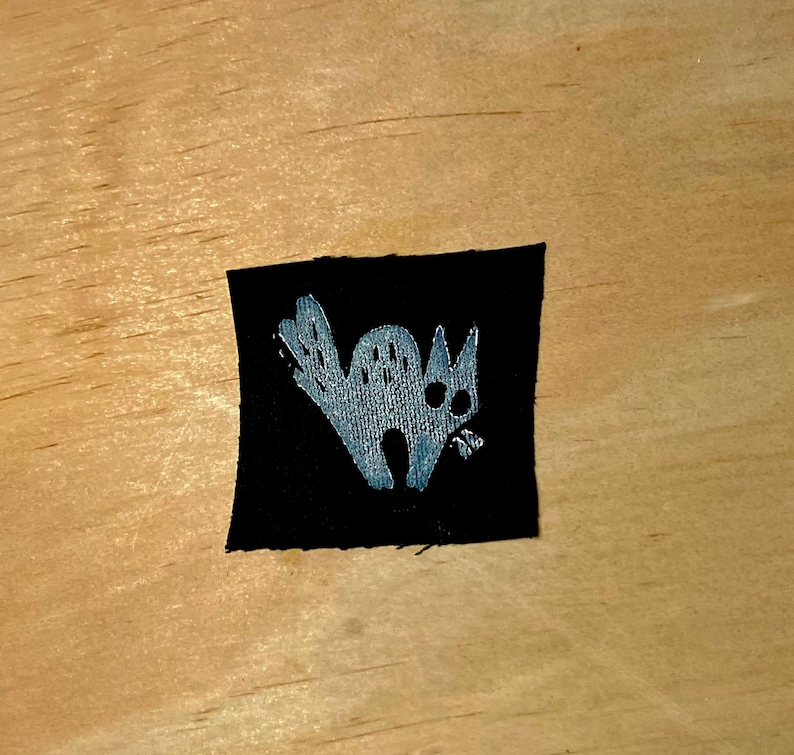 May include: A black square patch featuring a stylized, light gray cat illustration. The cat has large eyes and a curved tail, with a slightly open mouth. The patch is likely for clothing or accessories.