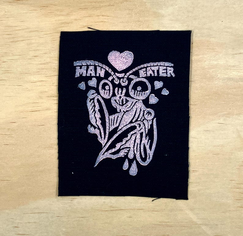 May include: Black fabric patch with a silver design and the words "MAN EATER". The design features a stylized insect with a heart above it and several small hearts around the insect. The patch is rectangular.