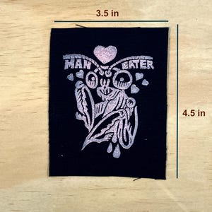 May include: A black patch with a silver design and the words "MAN EATER". The design includes a heart, a stylized face, and other decorative elements. The patch measures 3.5 inches by 4.5 inches.