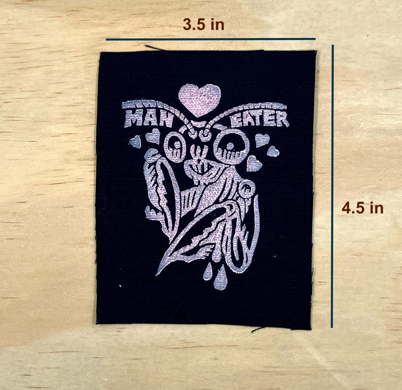 May include: A black patch with a silver design and the words "MAN EATER". The design includes a heart, a stylized face, and other decorative elements. The patch measures 3.5 inches by 4.5 inches.