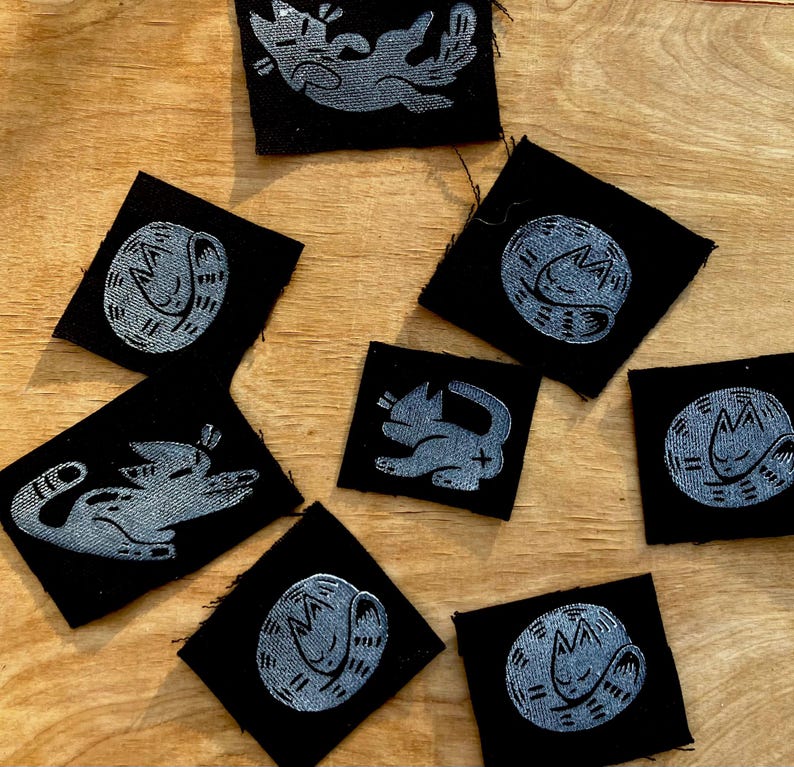 May include: A collection of black fabric patches with a silver cat design. The cat illustrations vary, showing different poses, including curled up and playful positions. The patches are square and arranged on a wooden surface.