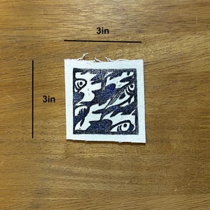 May include: A square, 3-inch by 3-inch, white fabric patch with a dark blue abstract design. The design features swirling shapes and eye-like details. The patch is placed on a wooden surface.