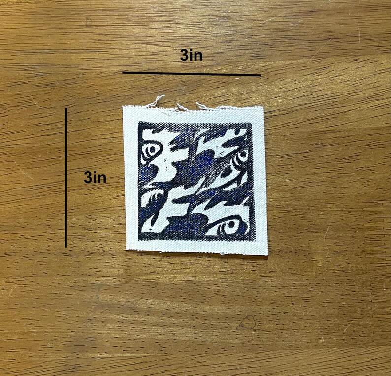 May include: A square, 3-inch by 3-inch, white fabric patch with a dark blue abstract design. The design features swirling shapes and eye-like details. The patch is placed on a wooden surface.