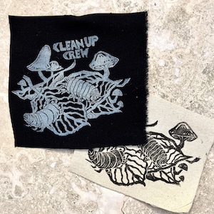 Puede incluir: Dos parches de tela, uno negro y otro blanquecino, con un diseño gráfico de setas estilizadas, un insecto segmentado y el texto "CLEAN UP CREW". El parche negro tiene un diseño azul claro, mientras que el parche blanquecino tiene un diseño negro.