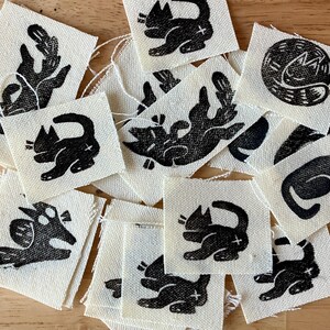 May include: A collection of small, square fabric patches with black cat illustrations. The cats are depicted in various poses, with a cross mark on their bodies. The patches are on a light beige background.