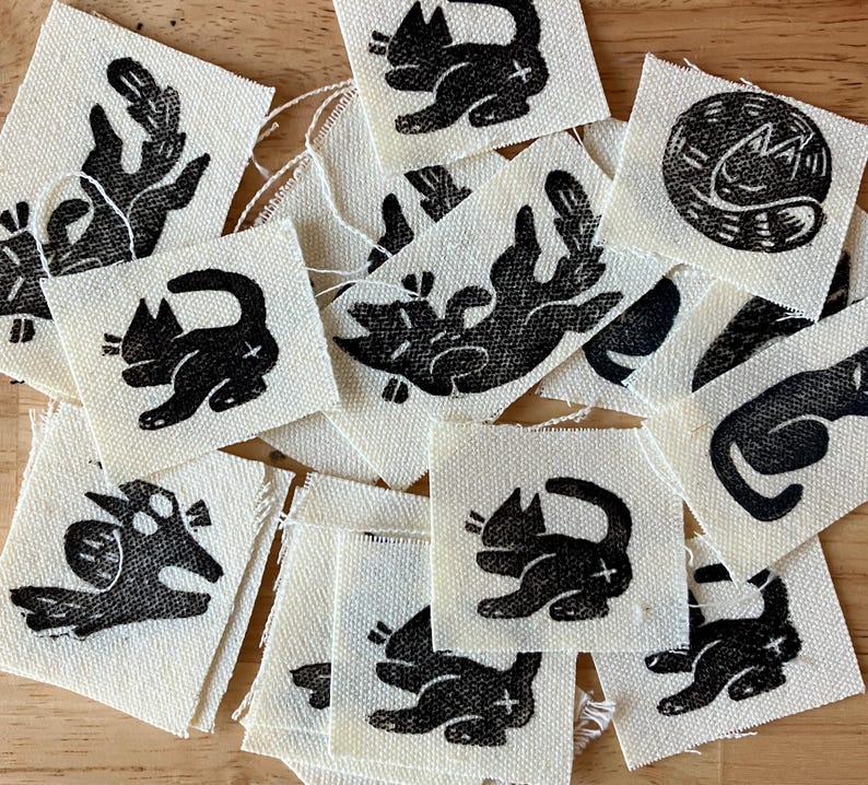 May include: A collection of small, square fabric patches with black cat illustrations. The cats are depicted in various poses, with a cross mark on their bodies. The patches are on a light beige background.