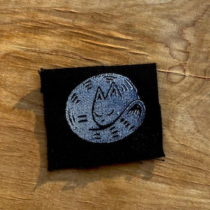 May include: A black fabric patch featuring a stylized, sleeping cat design in a light blue color. The cat is curled up in a circle, with simple lines indicating its features. The patch is square-shaped and has a stitched border.