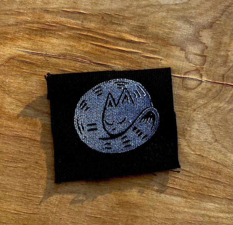 May include: A black fabric patch featuring a stylized, sleeping cat design in a light blue color. The cat is curled up in a circle, with simple lines indicating its features. The patch is square-shaped and has a stitched border.
