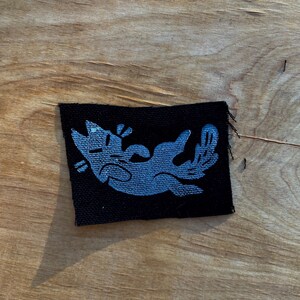 May include: A black patch featuring a light blue cartoon cat lying on its back with its paws in the air. The cat has a simple, stylized design. The patch is rectangular with frayed edges.