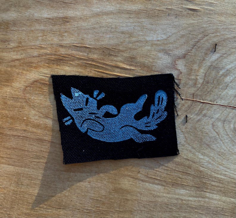 May include: A black patch featuring a light blue cartoon cat lying on its back with its paws in the air. The cat has a simple, stylized design. The patch is rectangular with frayed edges.