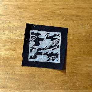 May include: A square, black fabric patch with a silver, abstract design. The design features swirling shapes and eye-like motifs within a silver border. The patch is placed on a wooden surface.