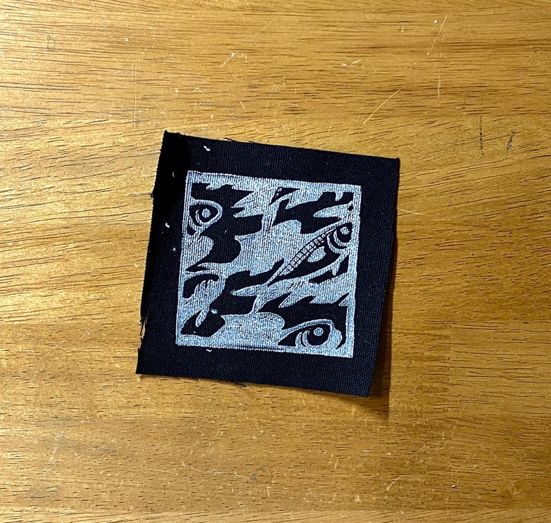 May include: A square, black fabric patch with a silver, abstract design. The design features swirling shapes and eye-like motifs within a silver border. The patch is placed on a wooden surface.
