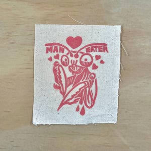 May include: A square, off-white fabric patch with a red graphic and text. The graphic features a stylized insect-like figure with a heart above it and smaller hearts around it. The text reads "MAN EATER".