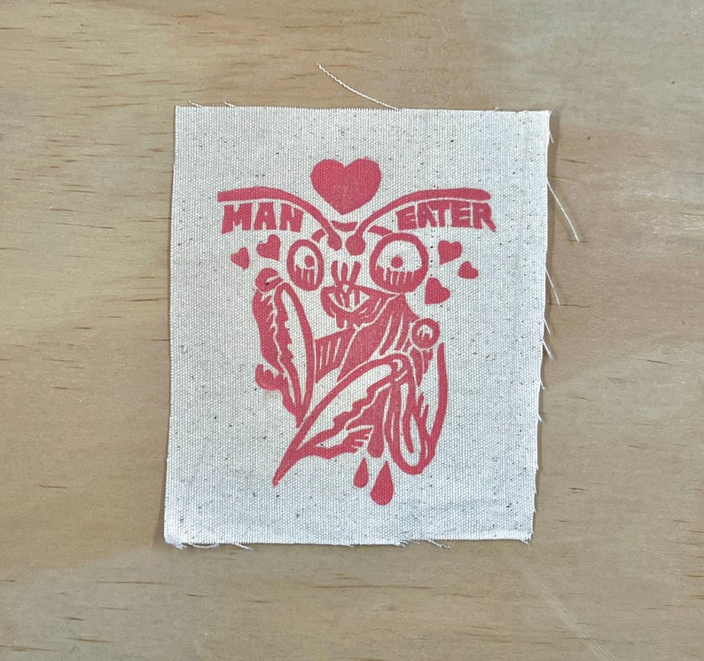 May include: A square, off-white fabric patch with a red graphic and text. The graphic features a stylized insect-like figure with a heart above it and smaller hearts around it. The text reads "MAN EATER".