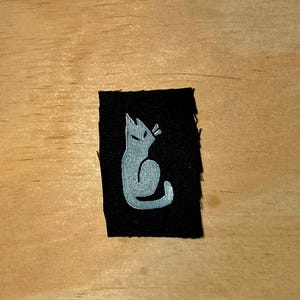 May include: A black fabric patch featuring a stylized silver cat silhouette. The cat is depicted in a seated position with pointed ears and a curved tail. The patch is rectangular with slightly frayed edges.
