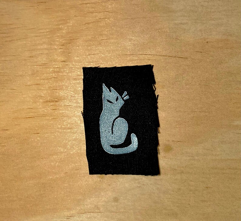 May include: A black fabric patch featuring a stylized silver cat silhouette. The cat is depicted in a seated position with pointed ears and a curved tail. The patch is rectangular with slightly frayed edges.