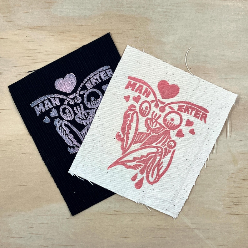 May include: Two fabric patches with the words "MAN EATER" and a graphic design. One patch is black with a light pink design, and the other is off-white with a red design. The patches are on a light brown wooden surface.