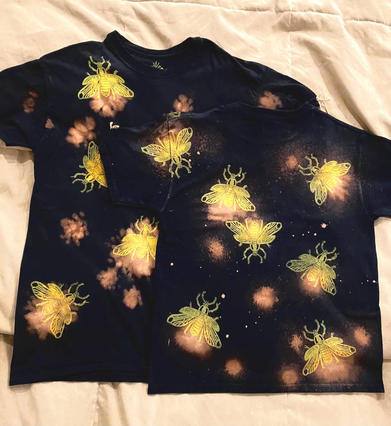 Op de afbeelding: Twee marineblauwe t-shirts met een gele insectenprint en een gebleekt, gespikkeld patroon. De insectenprint wordt herhaald op de shirts. De shirts hebben korte mouwen en lijken van katoen te zijn.