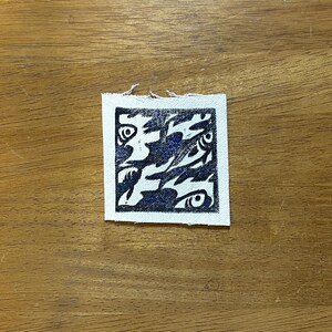 May include: A small, square fabric patch with a white border and a dark blue abstract design. The design features stylized shapes resembling eyes and other organic forms. The patch is placed on a wooden surface.