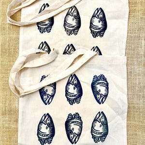 May include: Two cream-colored tote bags with long handles. Each bag features a repeating pattern of stylized, dark blue fish-like creatures. The bags are made of a sturdy fabric, suitable for carrying groceries or other items.