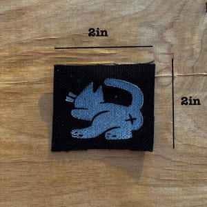 May include: A black square patch with a light blue cat design. The patch measures 2 inches by 2 inches. The cat is depicted in a playful pose with its tail curled. The background is a wooden surface.