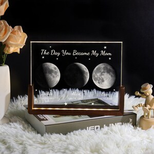 May include: A personalized moon phase night light with a wooden base. The black acrylic panel displays three moon phases, the text "The Day You Became My Mom", names, and dates. The light is on a white fluffy surface.