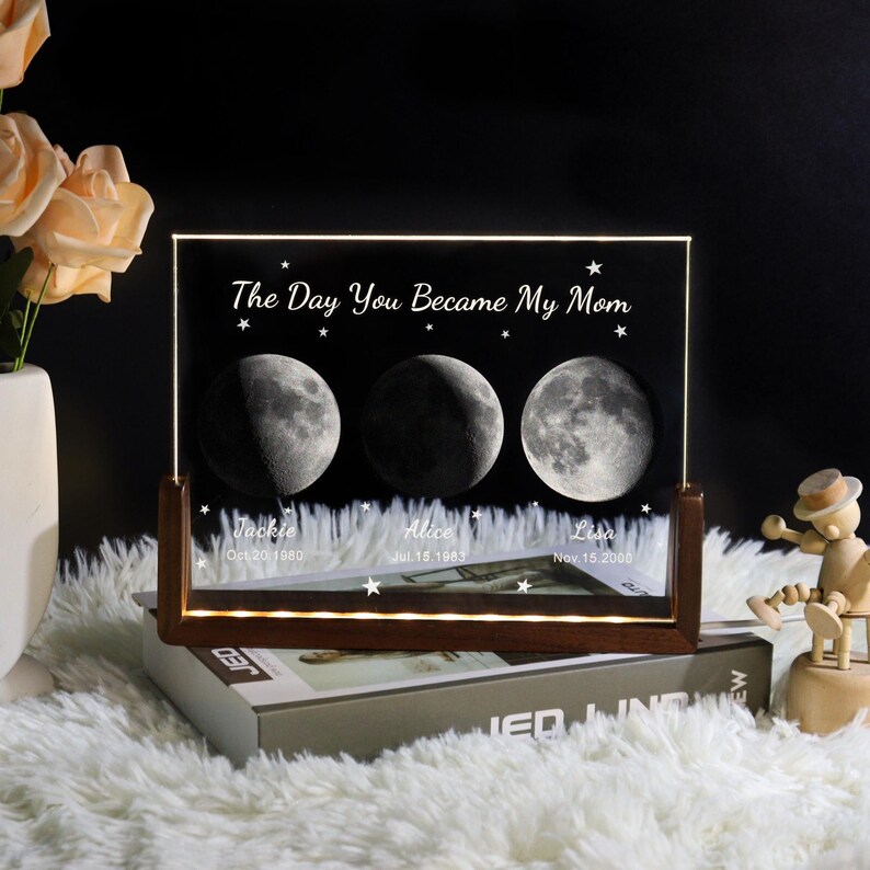 May include: A personalized moon phase night light with a wooden base. The black acrylic panel displays three moon phases, the text "The Day You Became My Mom", names, and dates. The light is on a white fluffy surface.