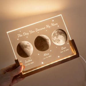 May include: A personalized acrylic night light with a wooden base. The light features three moon phase images and the text "The Day You Became My Mom." Names and dates are printed below each moon phase. The light is held by a hand.