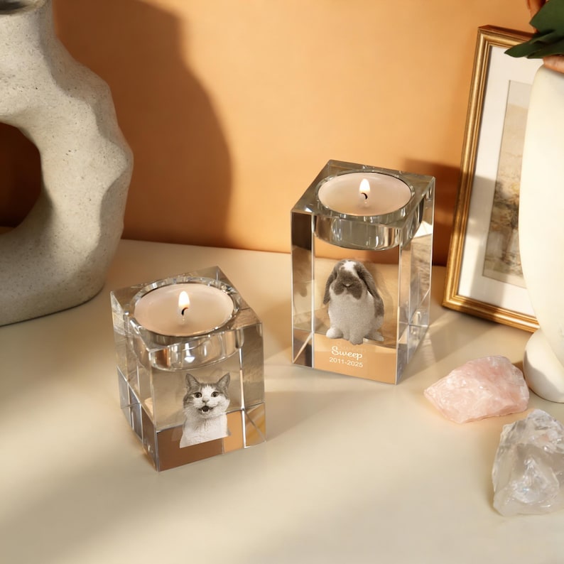 May include: Two clear glass candle holders with lit candles. One features a cat image, the other a bunny. The bunny candle holder has the text "Sweep 2011-2023". Decorative items for home decor.