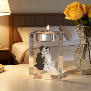 May include: A clear glass candle holder with a lit candle on top. The holder features a black and white photo of a couple kissing, with the text "Alex & Jane" below. Yellow roses in a glass vase are in the background.