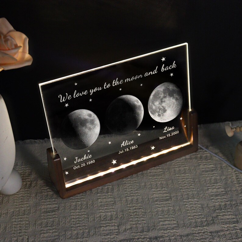 May include: A personalized night light with a wooden base and a clear acrylic panel. The panel features three moon phases, names, and dates, with the text "We love you to the moon and back". The light emits a warm glow.