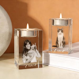 May include: Two clear glass candle holders with lit candles. One features a woman with a dog, the other a cat. The woman's holder has the name "Sophie" and paw prints. The cat's holder has the name "Felix" and paw prints.